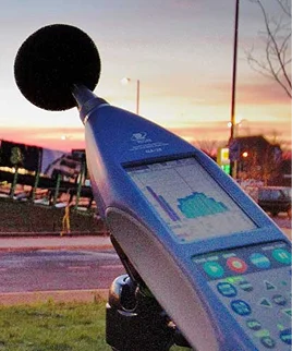 A sound level meter on a tripod, reading a level beside a road at dusk
